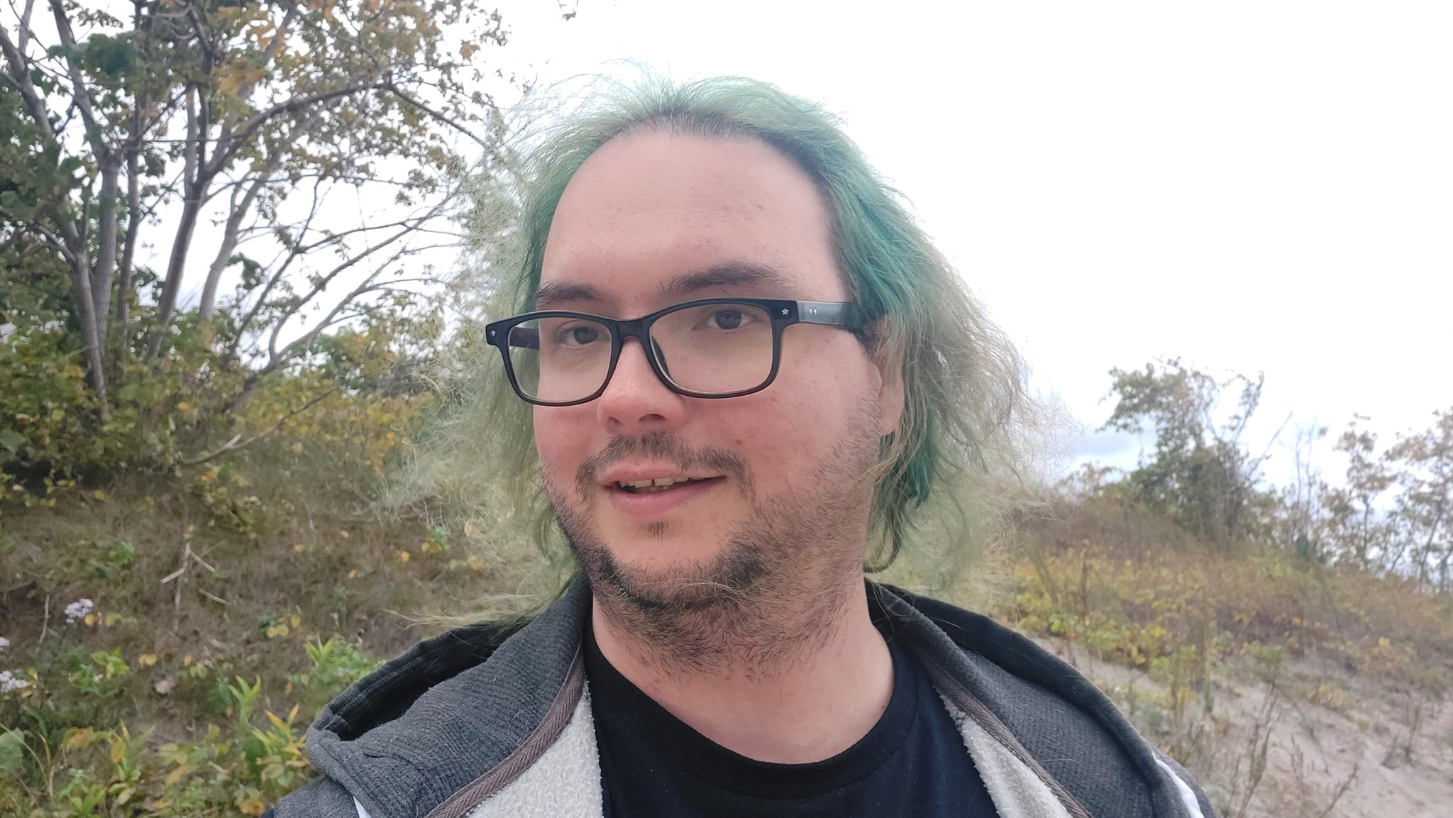 A man with long green hair stands on a grassy hill, the water is visible in the background